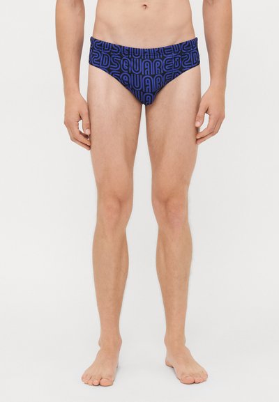 Modello maschile in piedi a piedi nudi, indossando slip da bagno blu con un audace motivo in lettere nere "Dsquared2" su sfondo bianco.