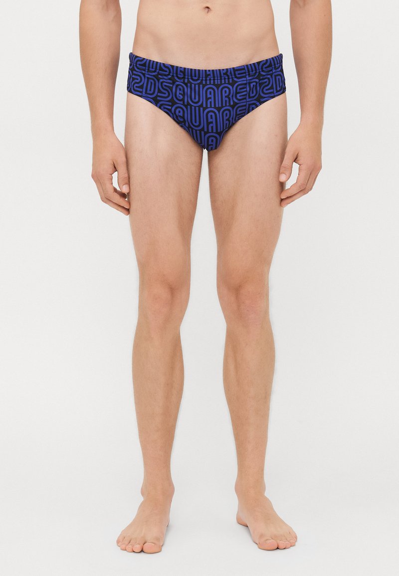 Männliches Model steht barfuß und trägt blaue Badehosen mit auffälligem schwarzen "Dsquared2" Schriftzug-Muster auf weißem Hintergrund.
