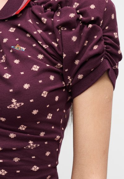 Lyhythihainen viininpunainen paita, jossa vaaleanpunainen kukkakuvio ja pieni kirjailtu logo rinnassa, päällä henkilöllä, jonka kyynärvarsi on näkyvissä.