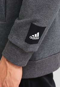 Sudadera gris con un acabado texturizado, que presenta un parche del logo negro de Adidas en la manga y puños acanalados. Detalles de costura visibles.