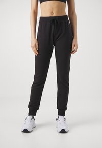 Black sweatpants with elastic waistband, adjustable drawstring, and ribbed cuffs. Smooth texture, tapered fit, paired with white sneakers.