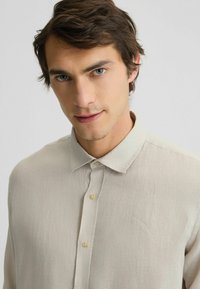 Camicia beige con bottoni, realizzata in tessuto testurizzato, con colletto morbido e bottoni piccoli. Design semplice senza motivi o accenti.