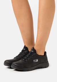 Zapatos deportivos negros con una parte superior de tela suave, sistema de cordones ajustable y una suela de goma texturizada con un logo prominente.
