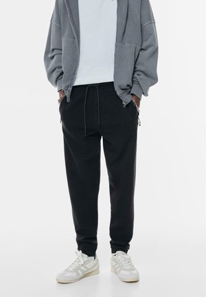Czarne spodnie dresowe z regulowanym sznurkiem, posiadające boczne kieszenie i luźny krój, zestawione z białymi i szarymi sneakersami.