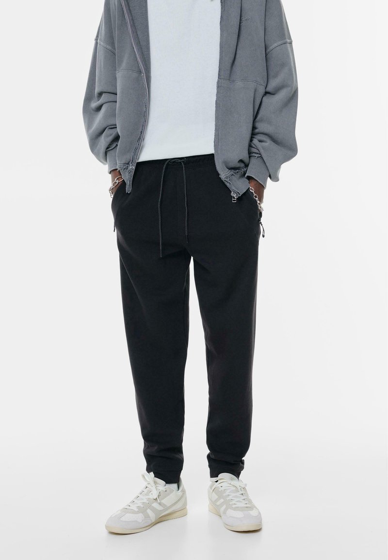 Pantaloni de trening negri cu șnur reglabil, având buzunare laterale și un croi relaxat, asortați cu adidași albi și gri.
