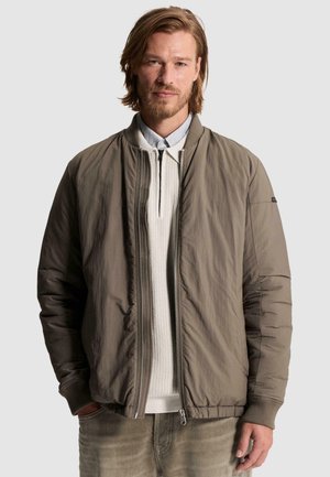 Homme aux cheveux châtain clair et à la barbe, portant une veste bomber gris-vert sur un pull blanc zippé et un pantalon clair.