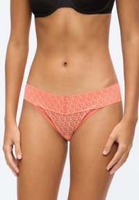 Donna che indossa slip bikini color corallo con motivo in pizzo e reggiseno nero, in piedi con le mani rilassate ai lati, davanti a uno sfondo uniforme.