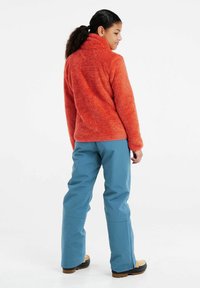 Fleece mikina v jasne oranžovej farbe s vysokým golierom, v kombinácii so svetlomodrými nepremokavými nohavicami. Hnedé topánky s textúrovanou podrážkou.