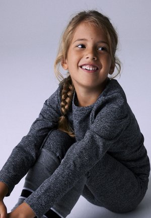 Sivi pleteni dukserica i odgovarajuće hlače. Teksturne, elasticne manžete; opušteni kroj. Dijete s pletenicama se smije dok sjedi.