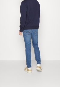 Homme portant un sweat-shirt bleu marine, un jean bleu et des baskets Veja beige, debout de dos face à la caméra sur un fond blanc uni.