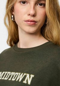 Jauna moteris su šviesia oda ir blondinėmis plaukais dėvi sidabrines lankų formos auskarus ir alyvuogių žalios spalvos džemperį su užrašu „MIDTOWN“.