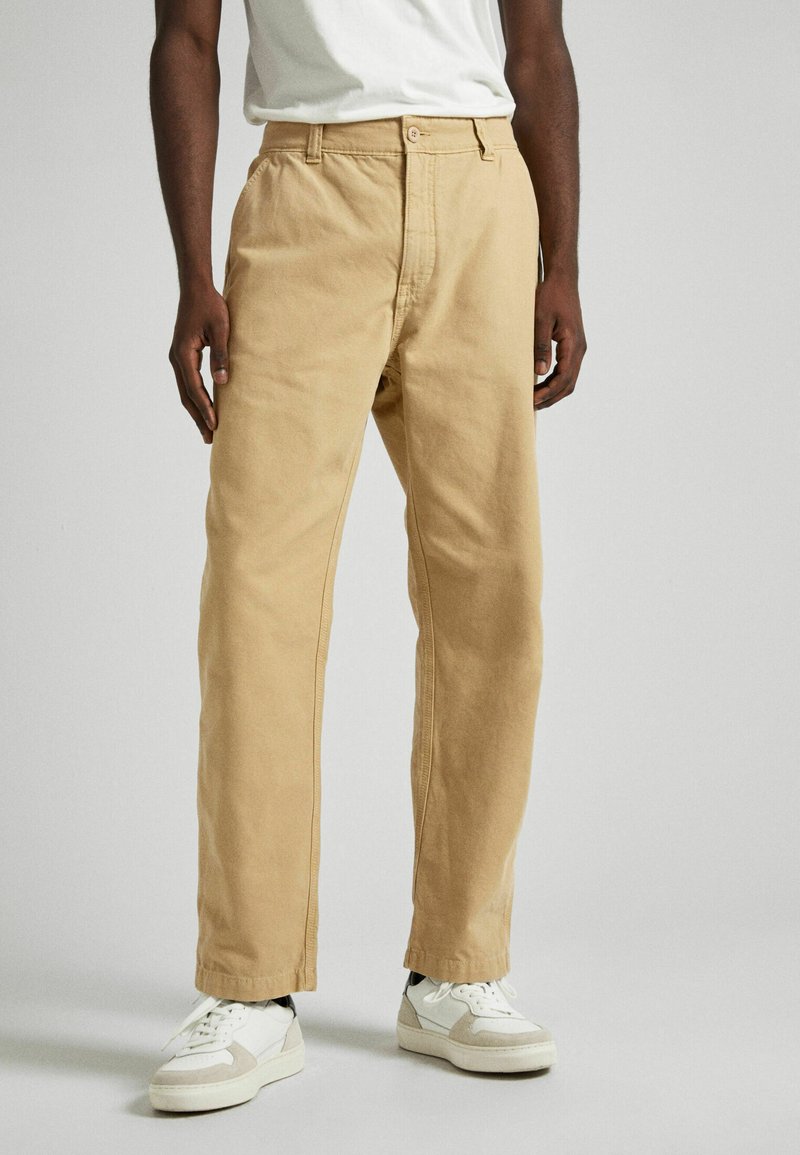 Šviesiai rudos medvilninės kelnės su tiesiu siluetu, su mygtukų uždarymu ir priekinėmis kišenėmis, derinamos su baltais sportbačiais. Glotnus tekstūras.