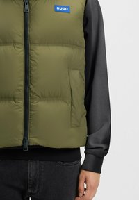 Chaleco acolchado verde oliva con cremallera frontal, logo azul destacado "HUGO" y bolsillos laterales, llevado sobre una sudadera oscura y vaqueros.