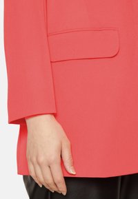 Blazer corallo con un design a taglio quadrato, spalla strutturata e una tasca a toppa visibile sul lato sinistro, realizzato in tessuto morbido.