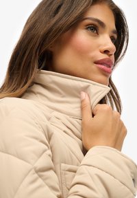 Beige gesteppte Jacke mit hohem Kragen, glatter Textur und Knopfdetails. Die Hand des Models hält sanft den Kragen und hebt das Design und die Passform hervor.