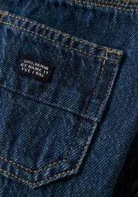 Denim fabric in deep blue color with contrasting tan stitching; features a rectangular back pocket with a black label reading "ORG. DENIM BY NAME IT EST. 1996."