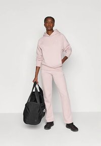 Light pink hoodie with a front pocket and matching sweatpants. Black duffel bag with sturdy handles. Model stands against a plain backdrop.