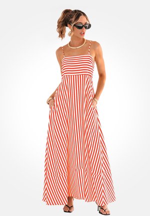 Femme portant une robe maxi sans manches rayée rouge et blanche, des lunettes de soleil noires, un collier en or et des sandales à talons noires, les mains dans les poches.