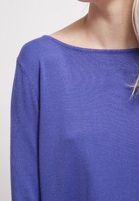 Fioletowy sweter dzianinowy z dekoltem w łódkę, o miękkiej teksturze i długich rękawach, charakteryzujący się delikatnym połyskiem oraz luźnym krojem.