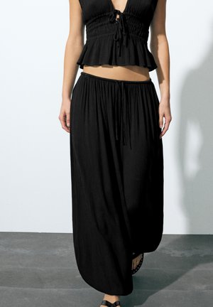 Žena, ktorá má na sebe voľný čierny bezrukávový crop top a široké čierne nohavice, stojí na šedej podlahe pred bielou stenou.