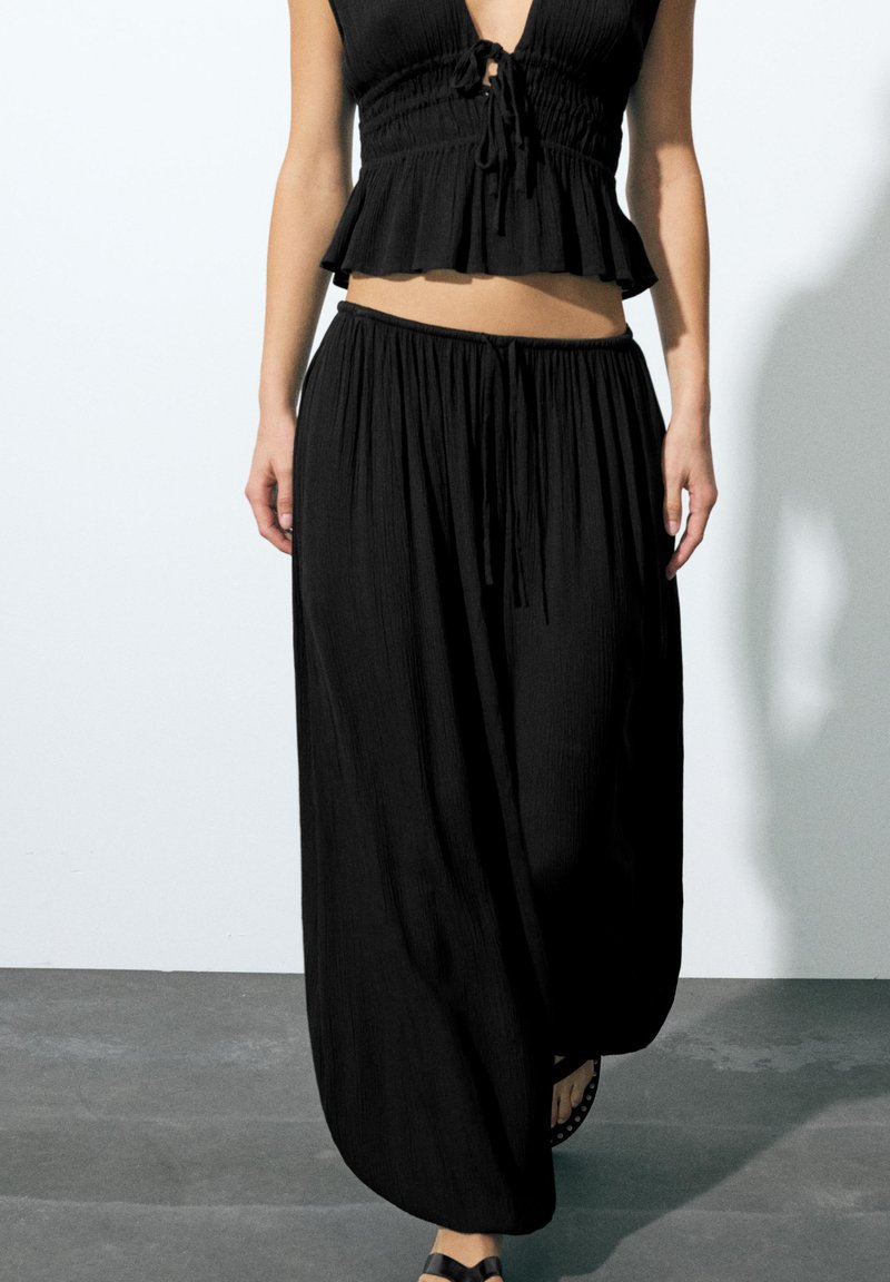 Femme portant un crop top noir ample sans manches et un pantalon noir à jambes larges, debout sur un sol gris contre un mur blanc.
