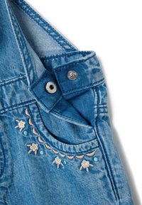 Denim tuinbroek in lichtblauw met geborduurd bloemenmotief. Voorzien van verstelbare bandjes en metalen drukknoopsluiting, met een zachte textuur en een vervaagde afwerking.