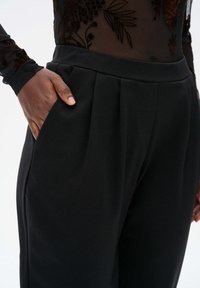 Zwarte geplooide broek met zijzakken, gemaakt van gladde stof. De tailleband zit net boven de heupen en heeft een strakke uitstraling zonder versieringen.