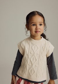 Cremefarbener Pullover aus gestricktem Stoff mit kurzen Ärmeln, der über einem dunklen Langarmshirt getragen wird und Kabelmuster sowie erhabene Texturen aufweist.