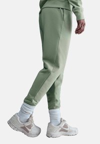 Pantaloni della tuta green in cotone con tasche laterali, gambe affusolate e vita con cordino, abbinati a scarpe da ginnastica bianche e beige e calzini bianchi.
