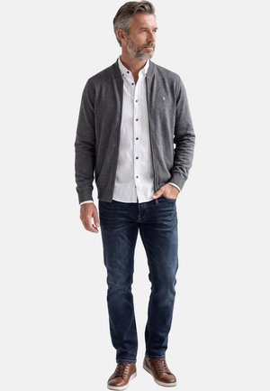 Uomo con capelli grigi che indossa un cardigan grigio con cerniera, camicia bianca con bottoni, jeans blu scuro e sneakers in pelle marrone, in piedi con una mano in tasca.