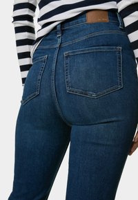 Jeans de mezclilla azul oscuro con un diseño de tiro medio, dos bolsillos traseros y un parche de cuero marrón en la cinturilla. Textura suave.