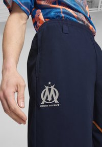 Pantalons de sport navy avec une taille élastique confortable, arborant un emblème argenté et le texte "DROIT AU BUT" sur la cuisse gauche.