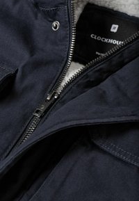 Veste bleu marine avec fermeture éclair métallique visible partiellement ouverte, bouton-pression près du col et doublure polaire à l'intérieur.