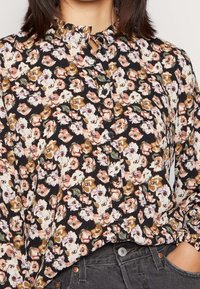 Femme portant une blouse noire à boutons avec un motif floral multicolore, rentrée dans un jean gris foncé taille haute.