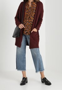 Burgundy lång cardigan över en brun leopardmönstrad blus, parat med avkortade, fransiga blå jeans och svarta slip-on skor. Svart crossbody-väska.