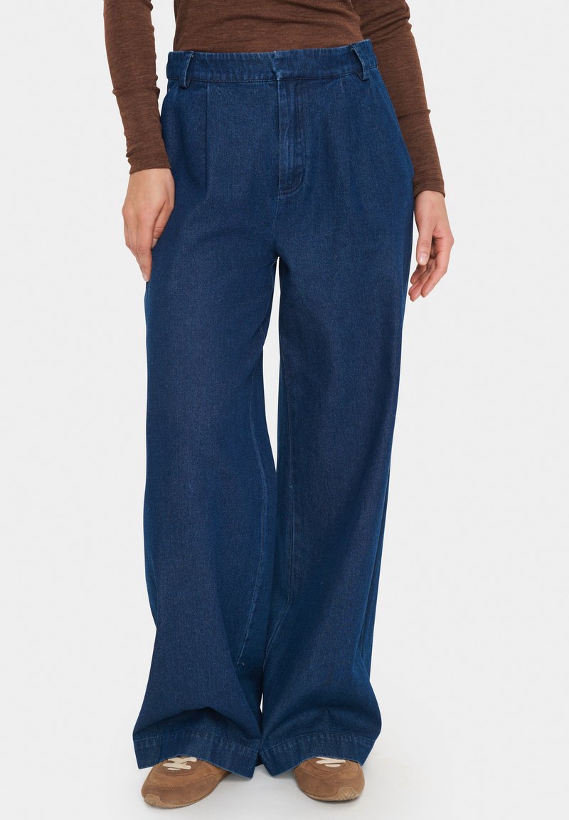 Vidbenade blå denimbyxor med två framveck, hög midja och en jämn textur, matchade med bruna skor.