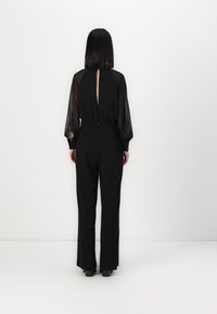 Zwart jumpsuit met doorschijnende lange mouwen, hoge hals en een sleutelgatdetail op de rug. Heeft een aansluitende taille en wijde pijpen.