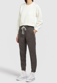 Persoon in een witte cropped sweatshirt, donkergrijze joggerpants met koord, en witte hoge sneakers, staand met één hand in de zak.