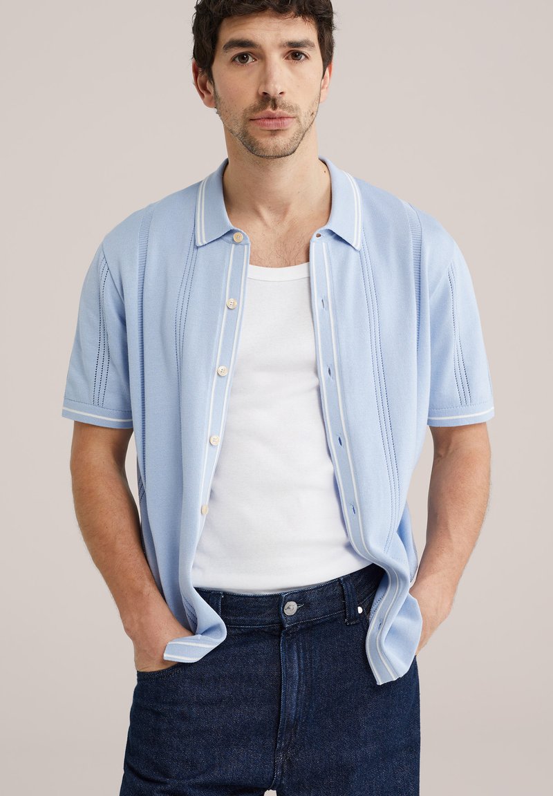 Uomo con camicia azzurra a maniche corte su canottiera bianca e jeans blu scuro, in piedi con le mani nelle tasche, sfondo neutro.