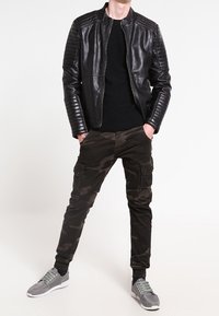 Mężczyzna w czarnej skórzanej kurtce, czarnej koszuli, spodniom w kamuflaż i szarych sneakersach, stojący z rękami w kieszeniach na białym tle.