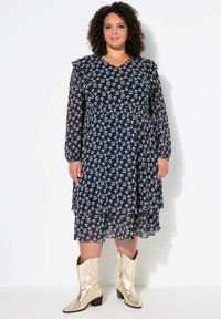 Vestido floral azul marino con mangas largas de tul, acentos con volantes, diseño en capas y una silueta fluida, combinado con botas de vaquero metálicas doradas.