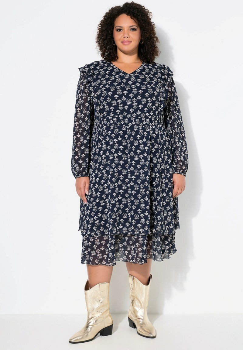 Vestido floral azul marino con mangas largas de tul, acentos con volantes, diseño en capas y una silueta fluida, combinado con botas de vaquero metálicas doradas.