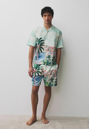 Young man standing barefoot wearing a matching short-sleeve shirt and shorts with a tropical beach and palm tree print.