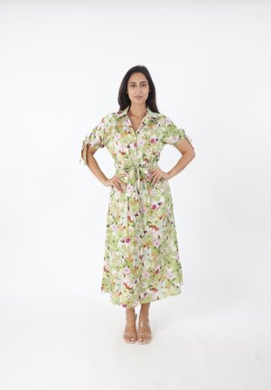 Kvinde stående med hænderne på hofterne iført en grøn blomsterprintet midi-kjole med opkørte ærmer og klare stroppestiletter mod en hvid baggrund.