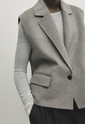 Person iført en grå ærmeløs uldblazer over en lysegrå langærmet top med sorte bukser, hånd i lommen.