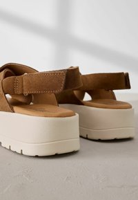 Sandali con plateau in suede marrone, cinturino regolabile e suola beige texturizzata, caratterizzati da una forma arrotondata e un sottopiede imbottito.