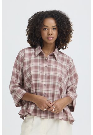 Göndörhajú nő laza rózsaszín és bézs kockás gombos ingben és világos színű nadrágban, egyszínű háttér előtt állva.