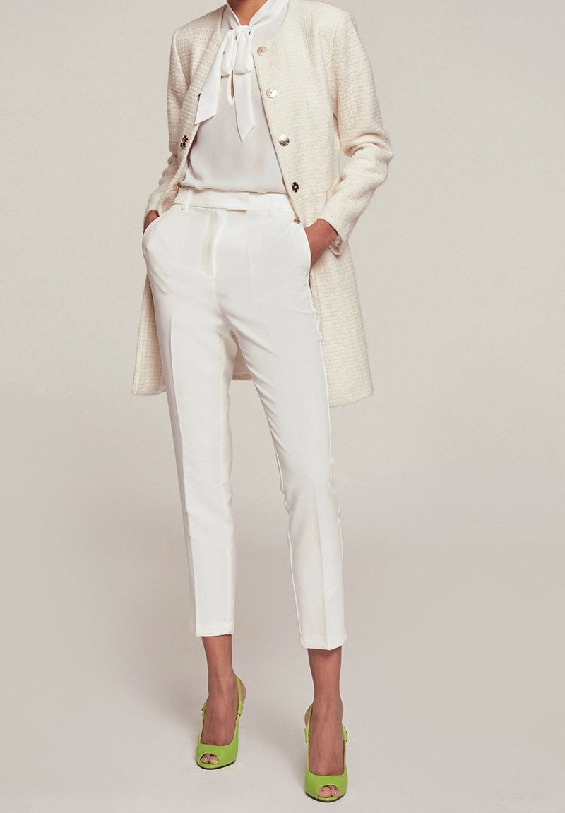 White tailored coat with gold buttons over a white blouse, paired with white tailored pants and bright green heels. Smooth textures throughout.