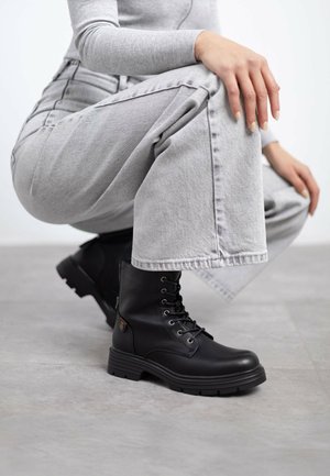 Botas negras de cordones hasta el tobillo con suela gruesa, acabado en piel suave y ojales de metal. Combinadas con unos jeans de cintura alta gris.