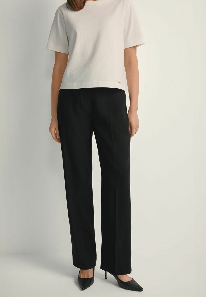 Kobieta ubrana w biały top z krótkim rękawem, czarne spodnie o prostych nogawkach oraz czarne wysokie obcasy w szpic, stojąca na tle jednolitego tła.